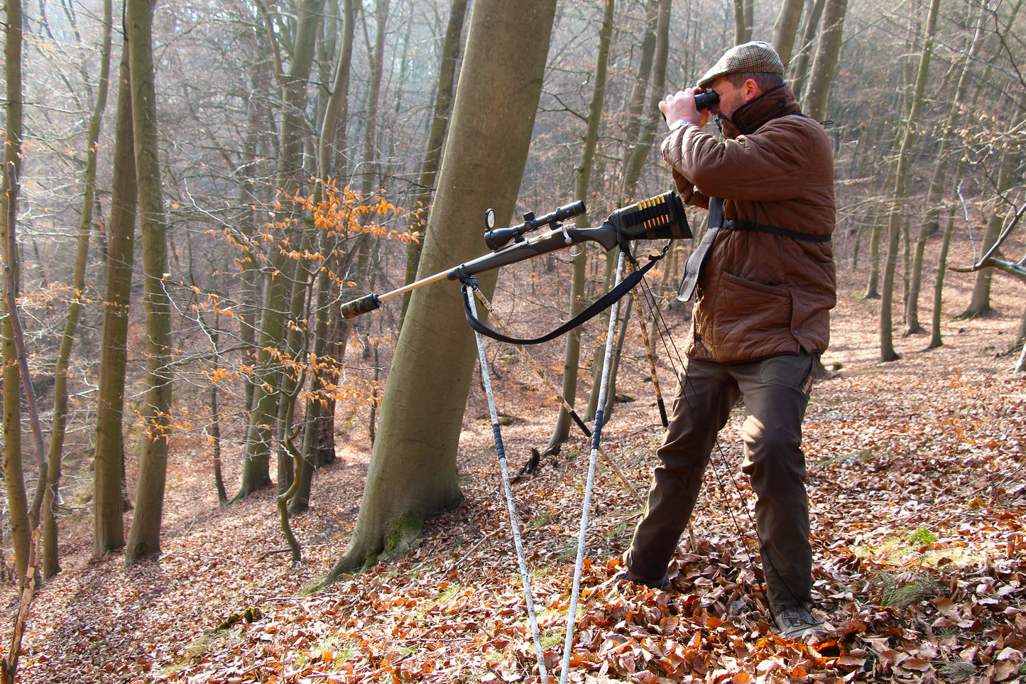 Ein Jäger steht in einem herbstlichen Laubwald und blickt durch ein Fernglas. Vor ihm ist ein Gewehr auf einem vierbeinigen Zielstock (Schießstock) aufgestellt, das zusätzlich mit einem Riemen gesichert ist. Die Waffe ist mit einem Schalldämpfer und Zielfernrohr ausgestattet. Der Jäger trägt eine braune Jacke, eine Schiebermütze und Jagdhose.