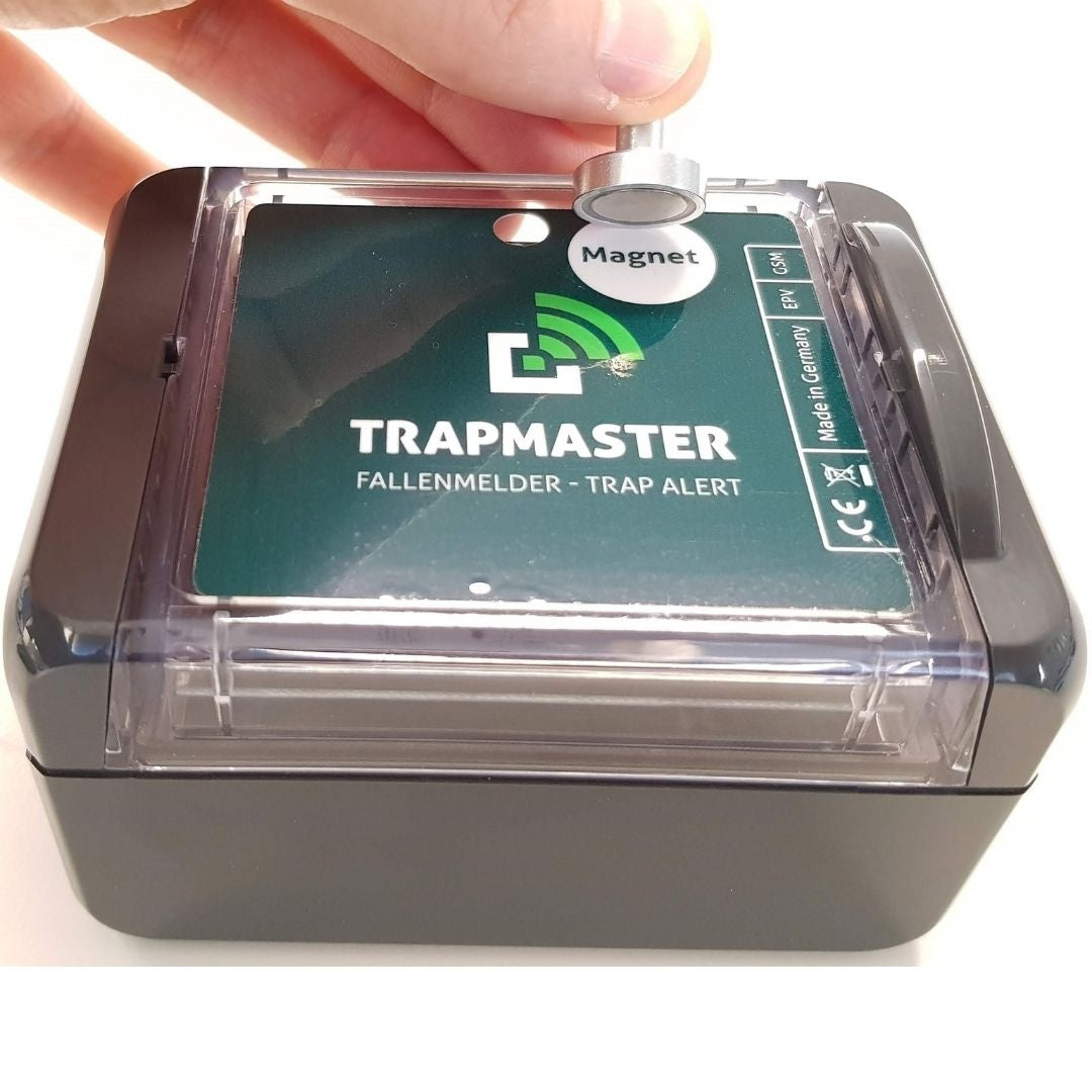 Eine Hand hält einen Magneten über einen elektronischen Fallenmelder des Typs Trapmaster. Der Magnet wird zur Auslösung oder Aktivierung des Geräts verwendet. Das grüne Bedienfeld mit Beschriftung und LED-Anzeige ist durch eine transparente Abdeckung geschützt.