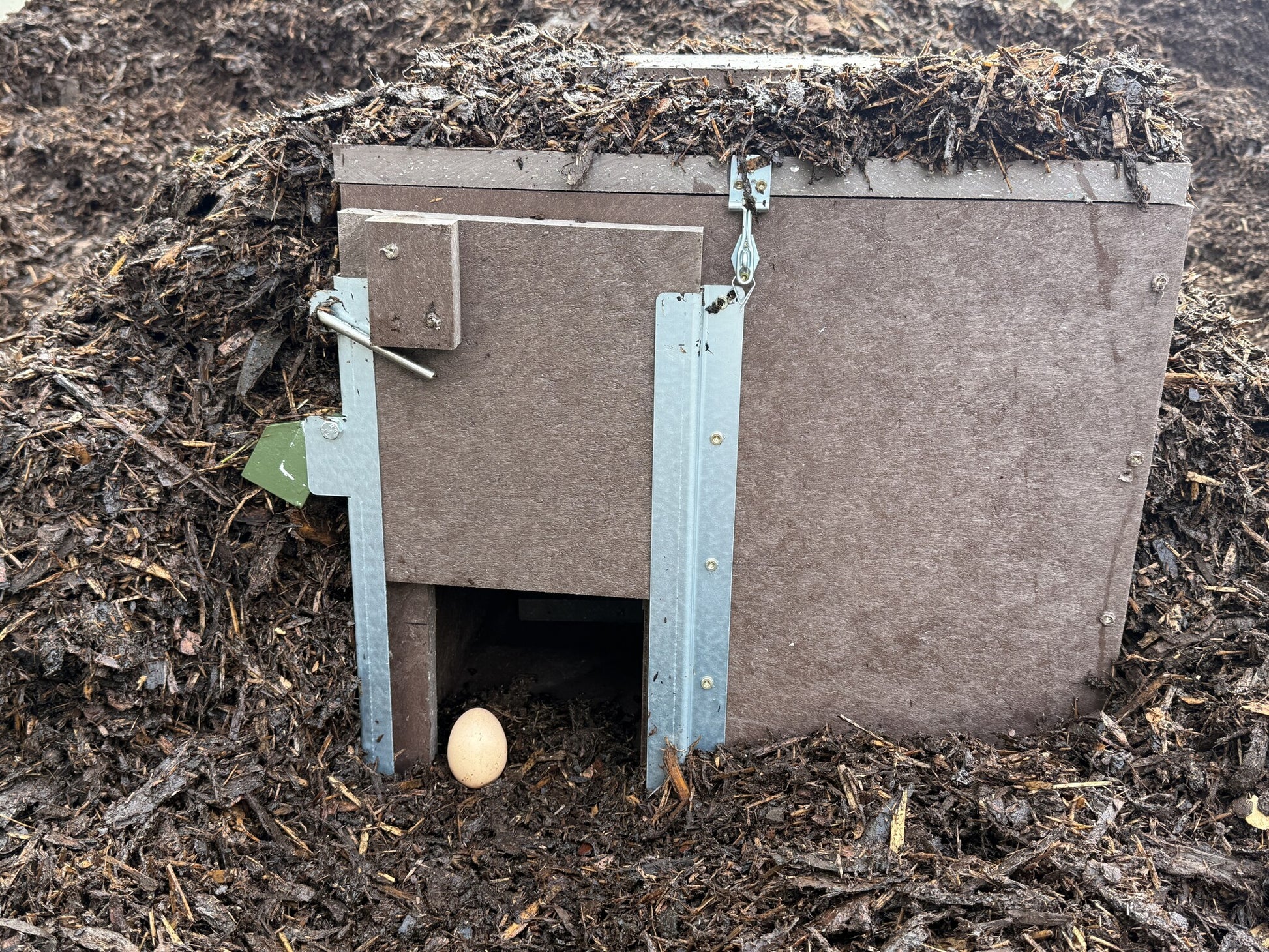 ie Abbildung zeigt eine kompakte Lebendfalle aus wetterfestem Verbundmaterial, aufgestellt in einem Haufen aus organischem Mulch oder Kompost. Die Falle ist mit seitlichen Verstärkungswinkeln aus Metall ausgestattet. Der Eingang ist bodennah freigelegt und mit einem braunen Ei als Lockmittel markiert. Die Oberseite der Falle ist mit Mulch bedeckt – zur Tarnung und besseren Wärmespeicherung. Diese Platzierung entspricht den Anforderungen für effektiven, tierschutzgerechten Lebendfang.
