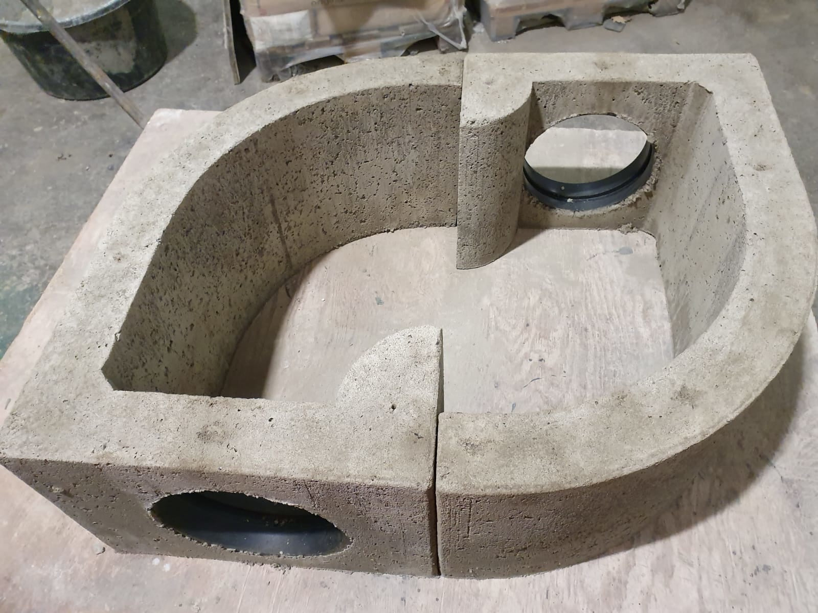 Auf dem Bild ist ein U-förmiges Betonbauteil mit zwei seitlichen Rohrdurchlässen zu sehen. Die Innenseite ist glatt gearbeitet, die Öffnungen sind mit eingelassenen Kunststoff-Rohraufnahmen versehen. Das Element ist in zwei Hälften gefertigt und dient vermutlich als Leitbogen oder Umkehrkammer für Lebendfangsysteme mit Betonrohrfallen. Es ermöglicht eine Richtungsänderung des Tierlaufs und die Integration von Fangtechnik im geschützten Inneren.