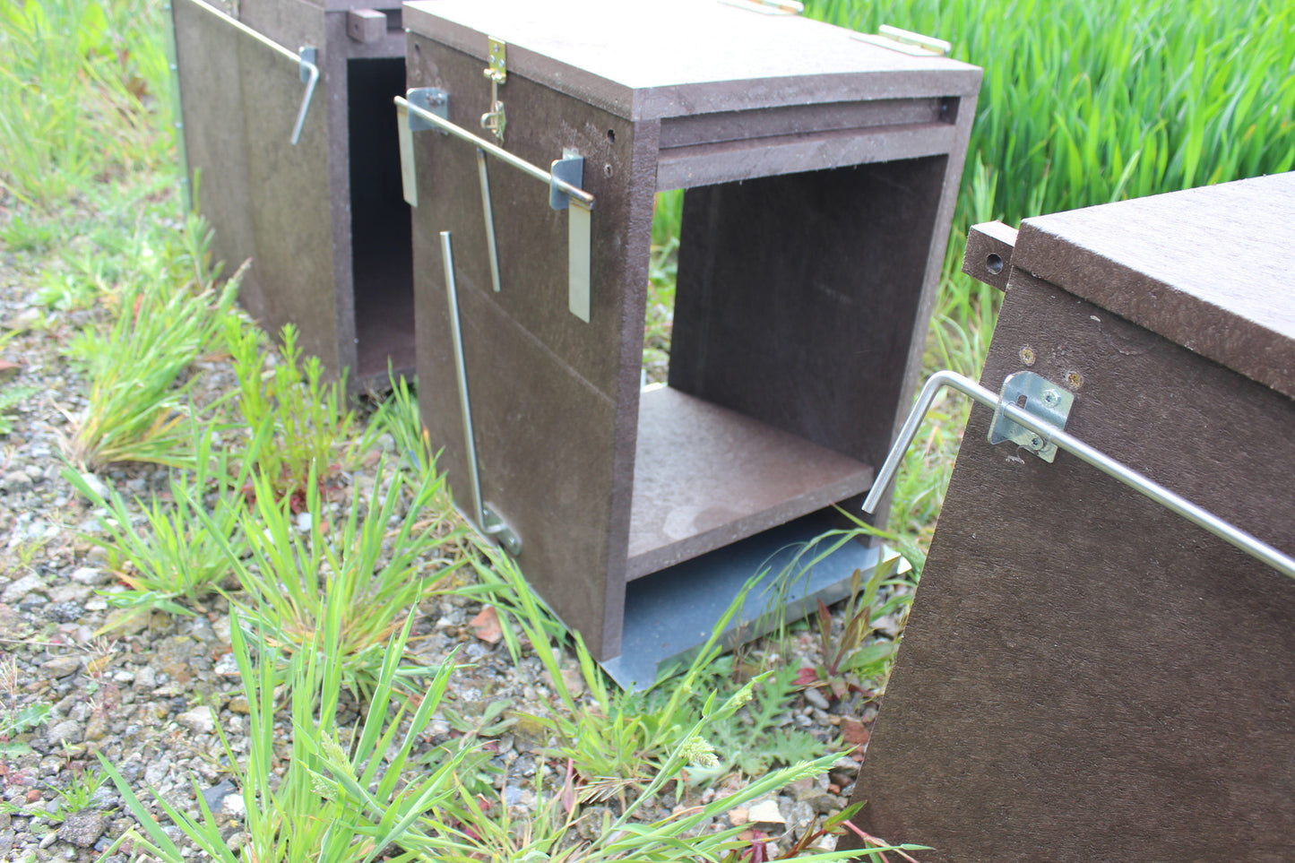 Auf dem Foto sind mehrere miteinander verbundene Fangkammern einer Lebendfalle in geöffneter Stellung zu sehen. Jede Kammer besteht aus wetterfestem Kunststoffmaterial mit Bodenführung und Fallstangenmechanik. Die Türen stehen offen, die Fallarme sind über außenliegende Metallbügel gesichert. Eine seitlich verlaufende Fangstange mit Laschen ist sichtbar. Die Aufstellung erfolgt auf Schotterboden direkt am Feldrand – optimal für Wechsel von Raubwild wie Fuchs oder Waschbär.