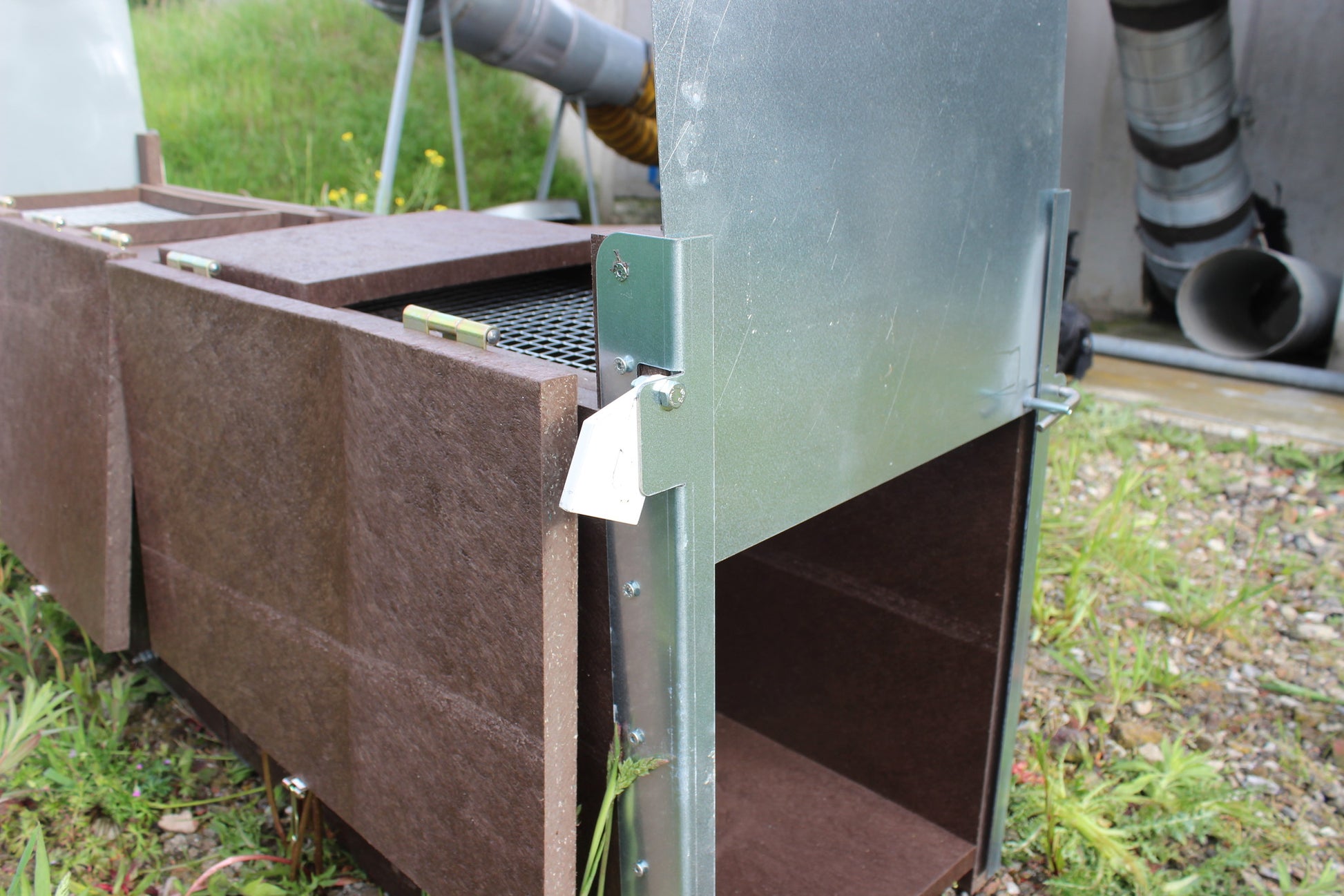 Auf dem Bild ist eine Lebendfalle in Nahaufnahme zu sehen. Die Seitenwände und der Korpus bestehen aus wetterfestem Recyclingmaterial in Holzoptik. Der Eingang der Falle ist mit einem stabilen verzinkten Metallrahmen verstärkt und verfügt über eine Schwenkklappe mit Arretierung. Im oberen Bereich sind Klappdeckel mit Metallscharnieren sichtbar – darunter vermutlich Gitter- oder Trittmechanik. Die Falle steht auf einem Naturboden mit Kies und Wiese, im Hintergrund technische Infrastruktur.