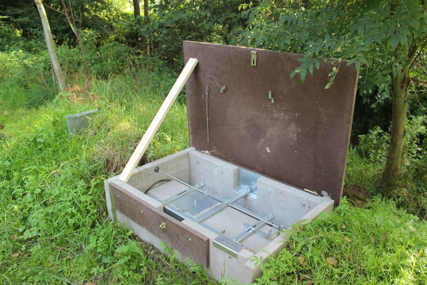 Geöffnete Raubwildfalle mit aufgeklappter brauner Abdeckung, die mit einem Holzstab abgestützt ist. Im Inneren sind metallische Auslösemechanismen und Führungsschienen sichtbar, eingebettet in einen rechteckigen Betonkörper. Die Falle befindet sich in einer bewachsenen Waldlichtung mit Gras und Büschen.