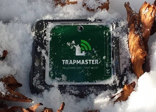 Ein Trapmaster-Fallenmelder liegt halb eingeschneit auf dem Waldboden, umgeben von trockenen Blättern. Das Gerät ist deutlich erkennbar mit grüner Oberfläche und weißem Logo.